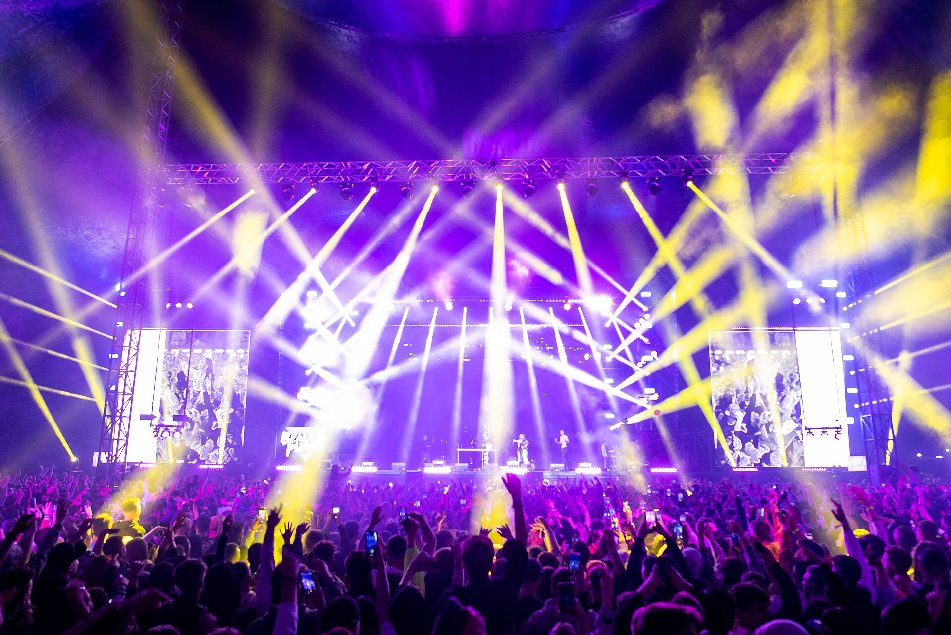 Crowd with hands raised before a brightly lit festival stage at night